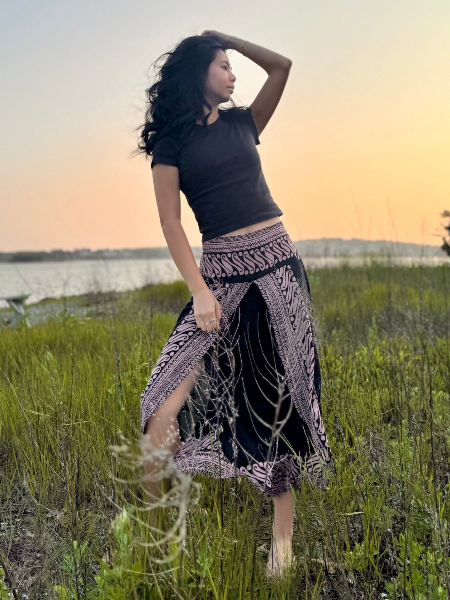 Model wearing black and pink patterned Hanna capri pants with front slits, standing in a grassy filed near the ocean.