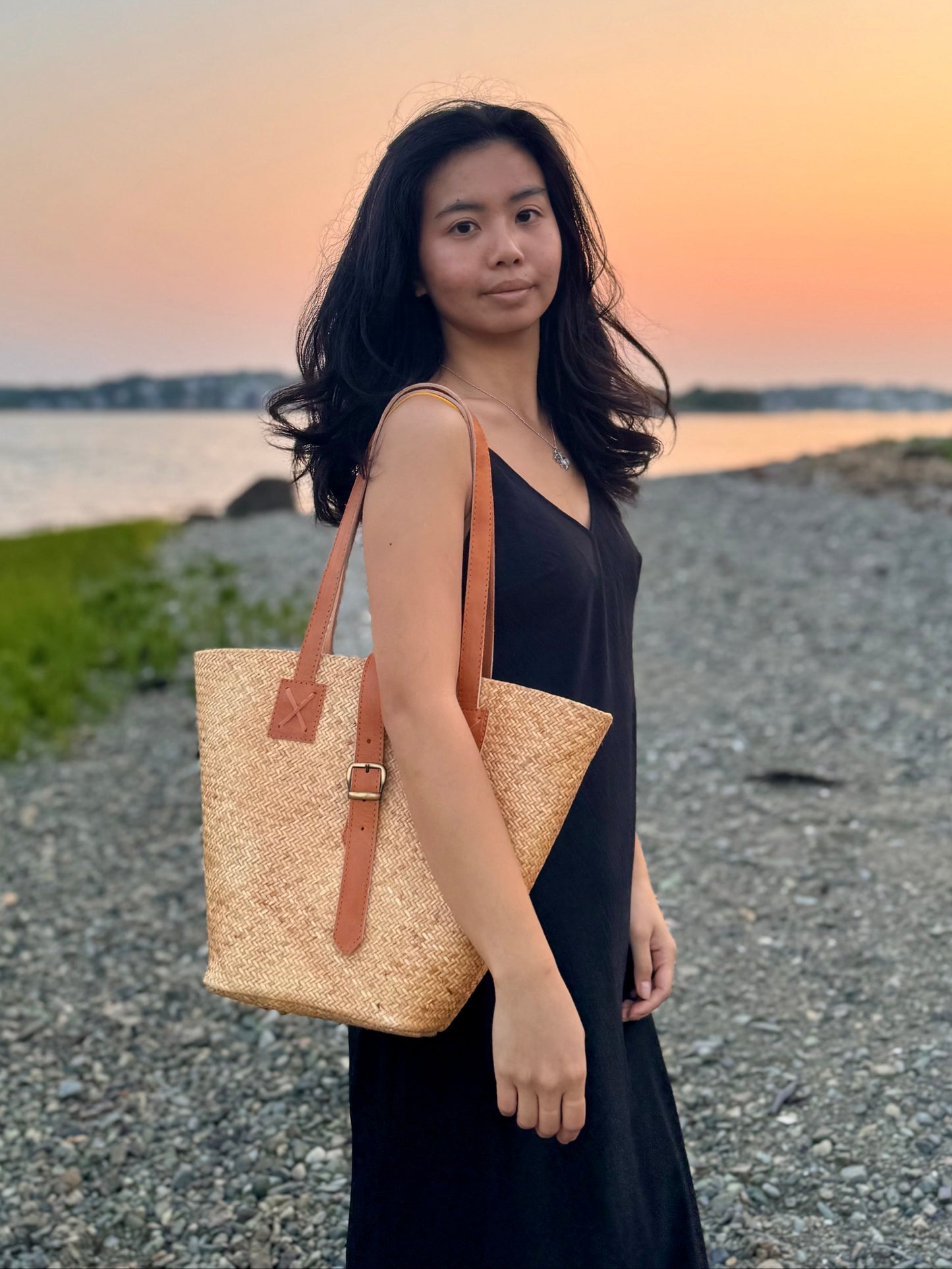 Model wearing Sari Beach bag over her shoulder, near the ocean during sunset.