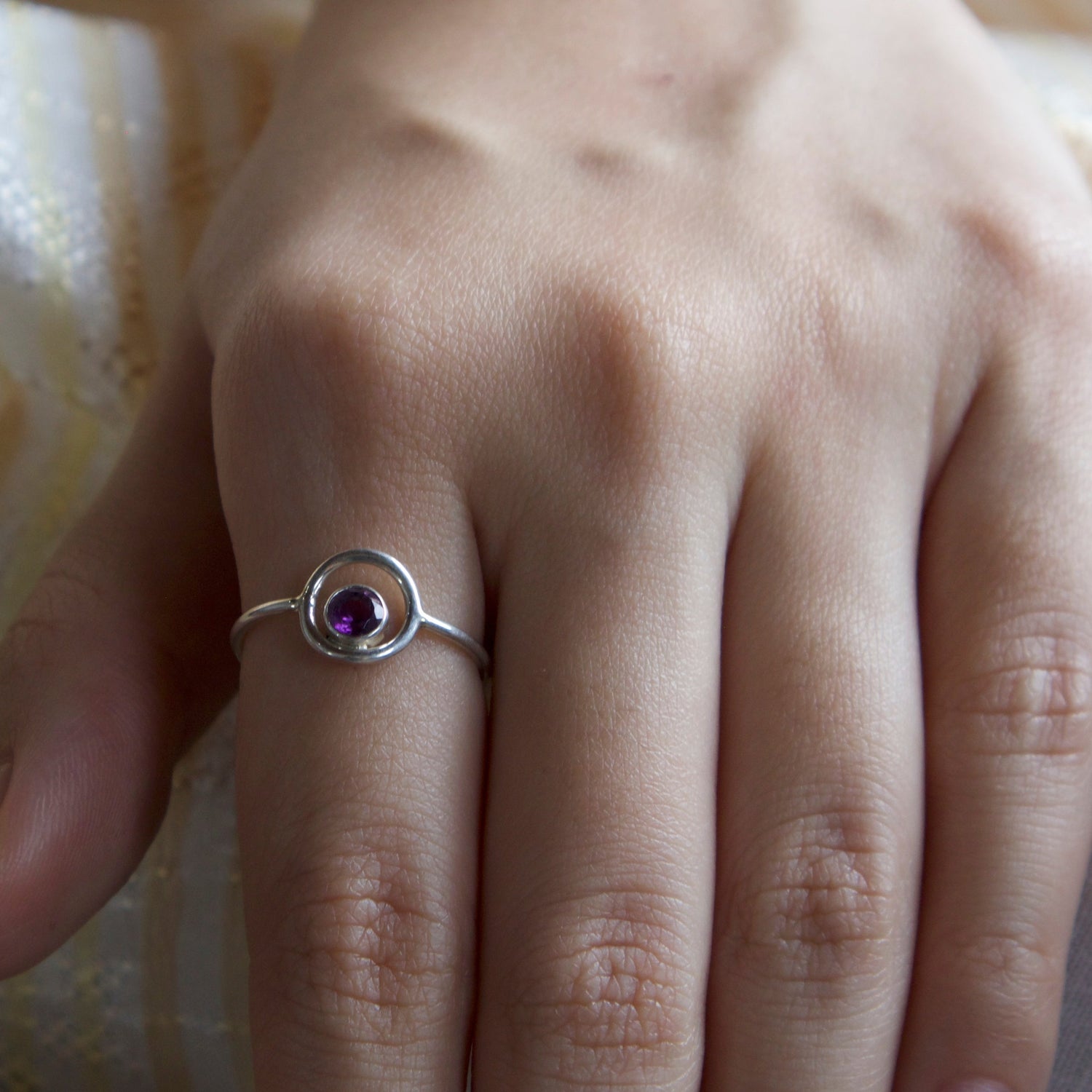 Hand wearing a silver ring with a purple gemstone on a textured fabric background
