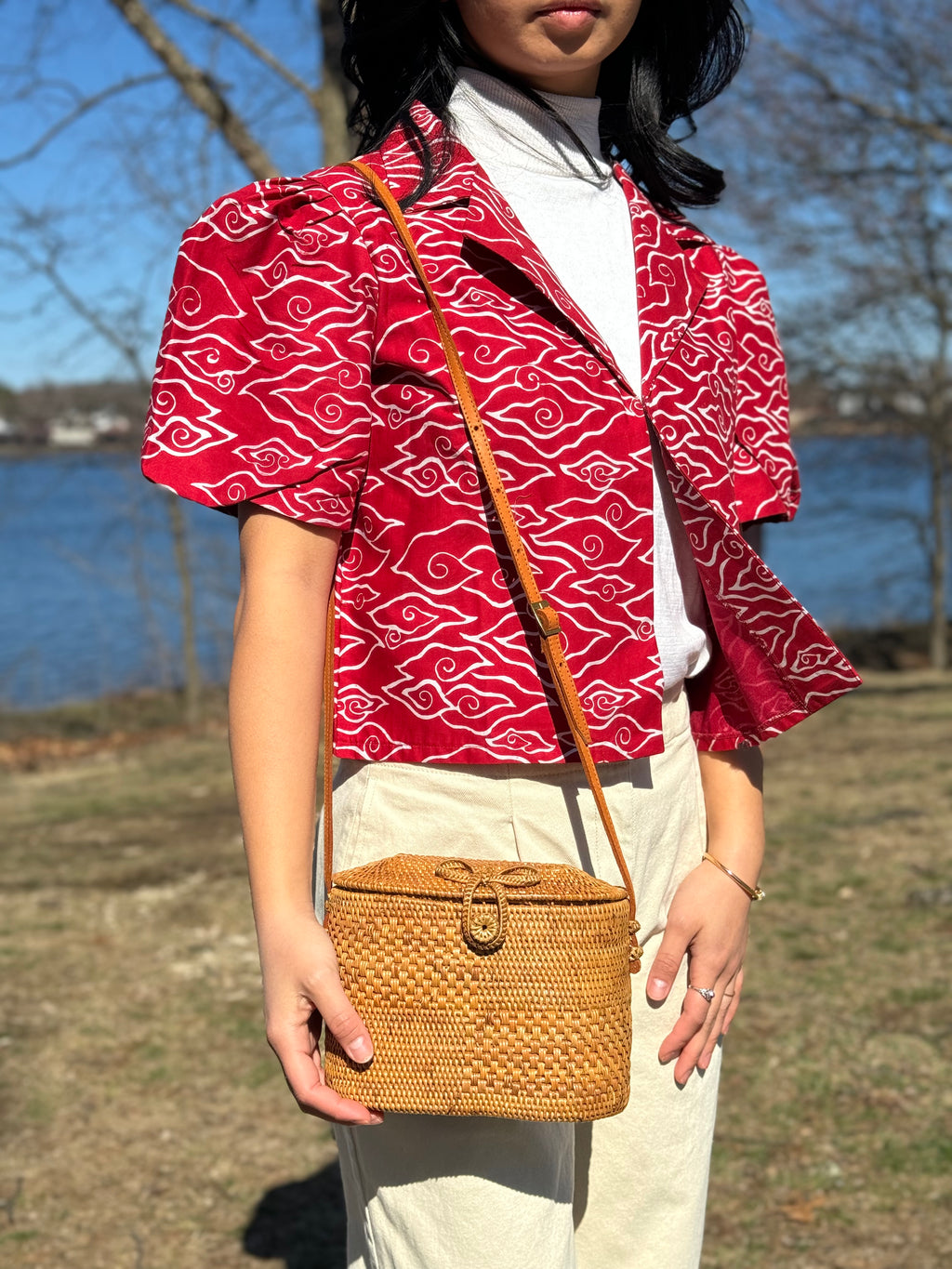 Person wearing a red patterned blouse holding a woven bag outdoors.