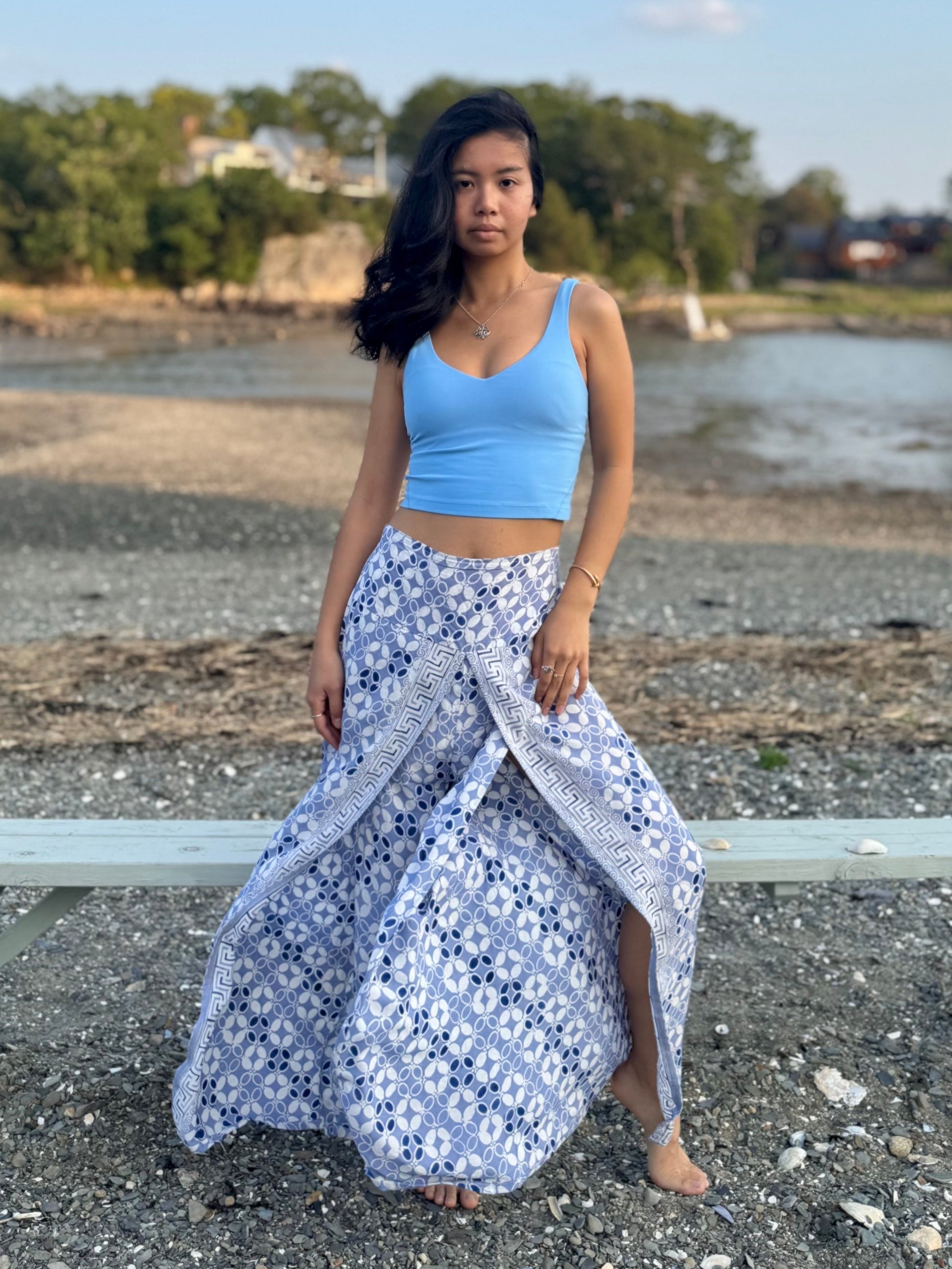 Model wearing high slit beach pants in blue and white print, standing on a rocky beach.