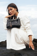 Kirana mini woven leather top handle bag in black with gold hardware and tassel detail, held by a woman in a white linen outfit against a coastal backdrop.