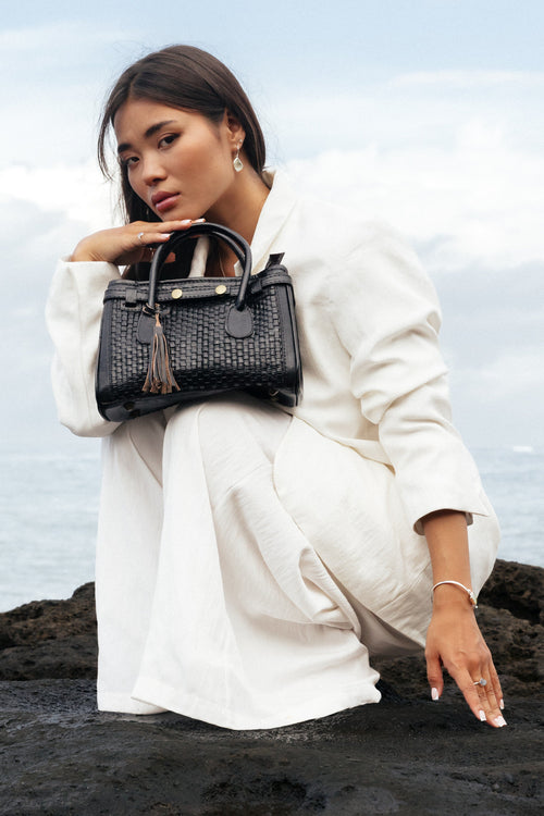 Kirana mini woven leather top handle bag in black with gold hardware and tassel detail, held by a woman in a white linen outfit against a coastal backdrop.