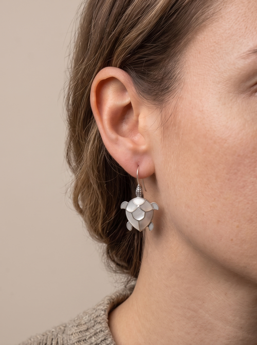 Close-up of a person wearing silver turtle-shaped earrings against a neutral background