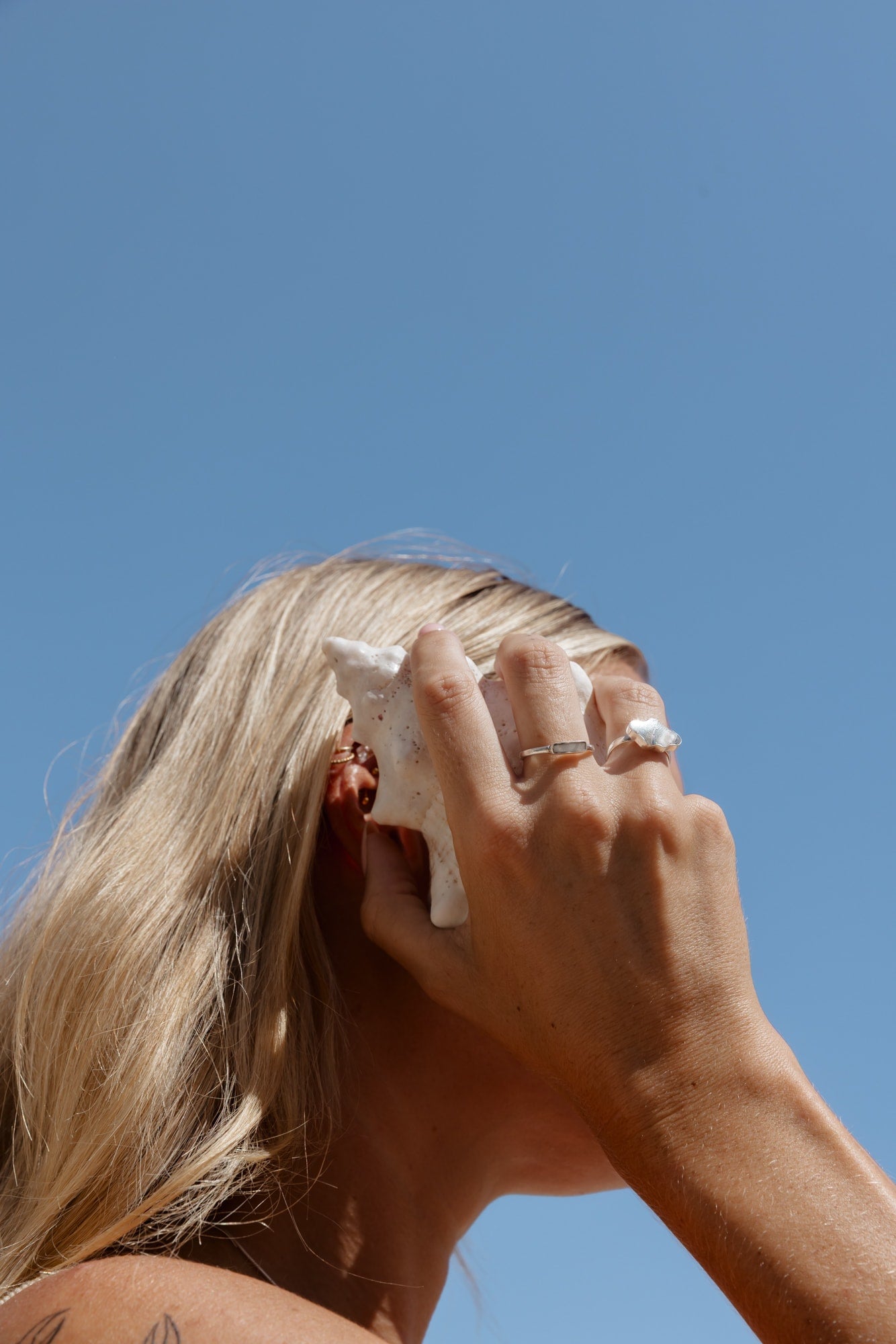 Model wearing shell ring holding a conch shell to her ear.